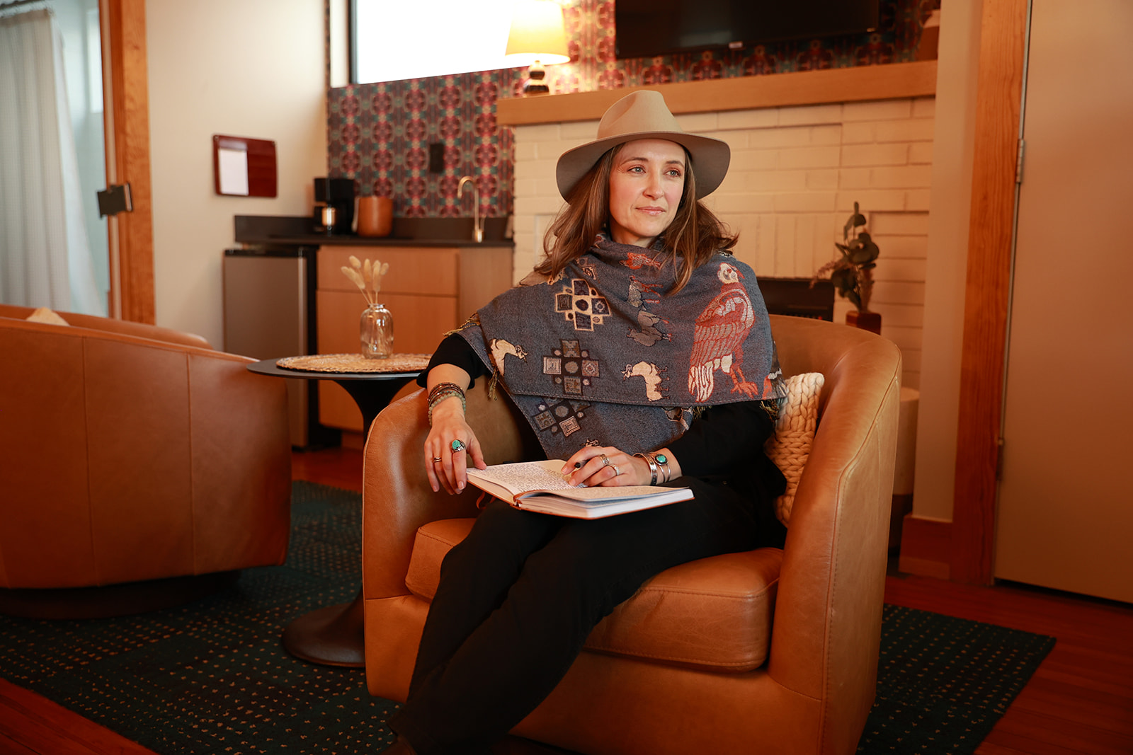 Amber, sitting in a leather chair, wearing a Peruvian shawl and a brown hat while journaling and gazing out. The background has a warm amber glow.
