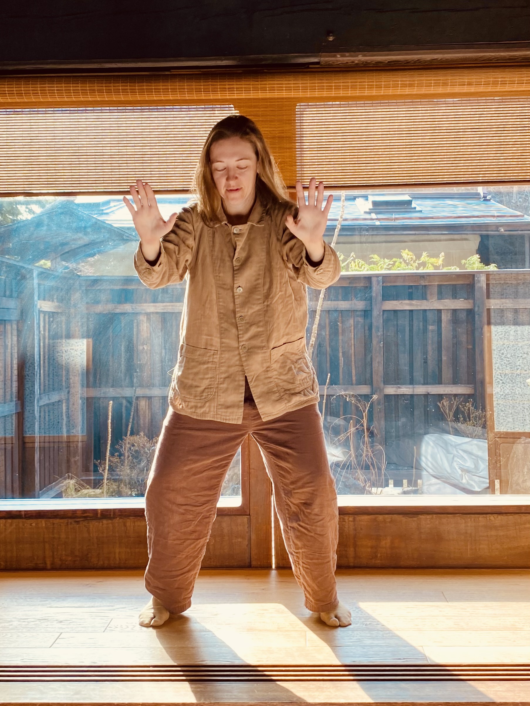 Amber is wearing a brown pajama set and practicing Qi Gong. The background includes a big window displaying a wooden fence and garden.