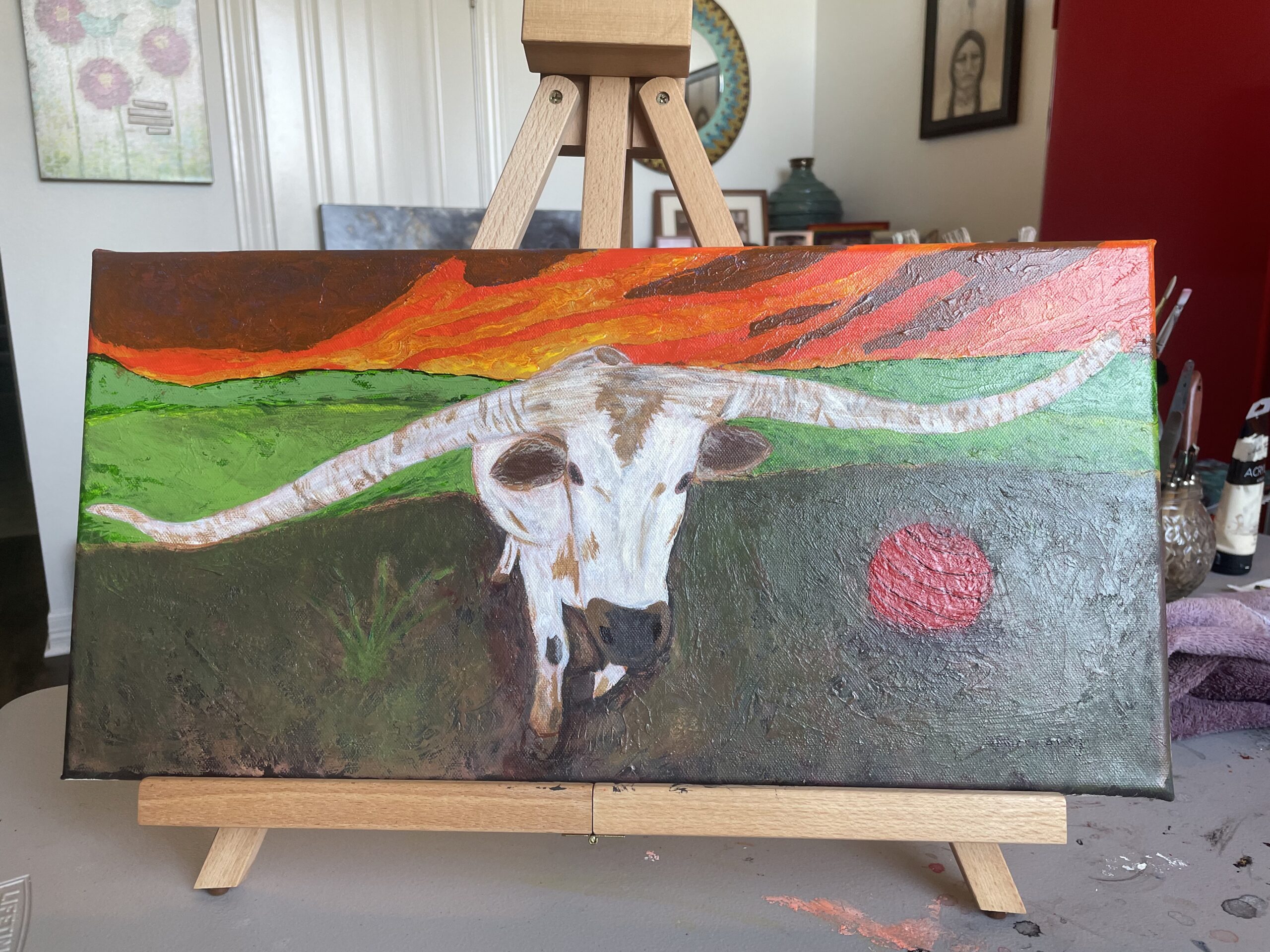 A painting of a big white longhorn with a red ball next to him in the dirt. There is a vivid sunset behind him.
