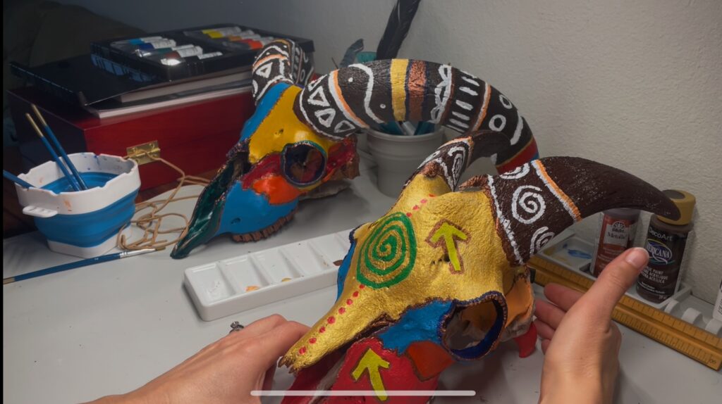hands are picking up the painted goat skull; the Aoudad skull is in the background on the desk