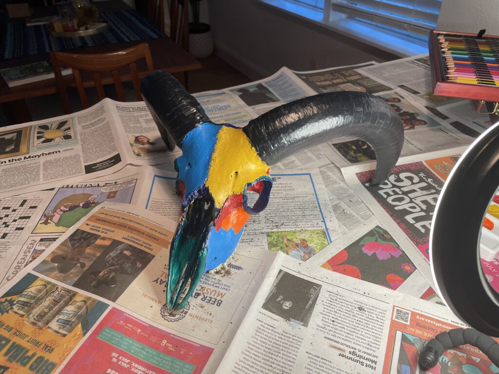 an Aoudad sheep skull with yellow, blue and orange colors on the face and black electrical tape wrapping the horns