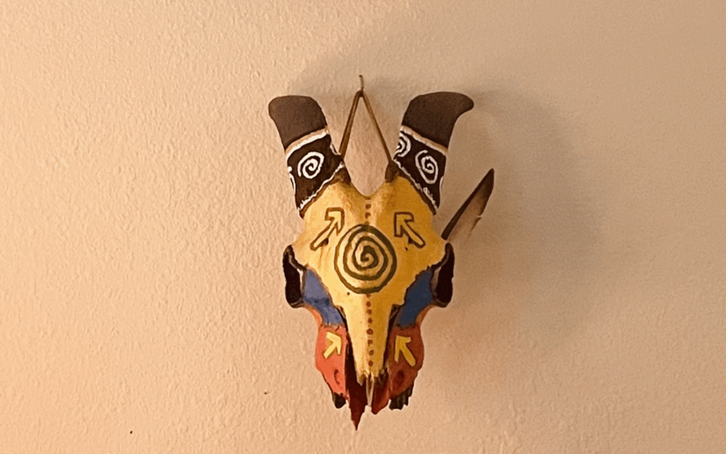 A painted goat skull with a feather stuck on the side; gold, pink, blue, brown and a light green pachamama symbol on the center of the forehead