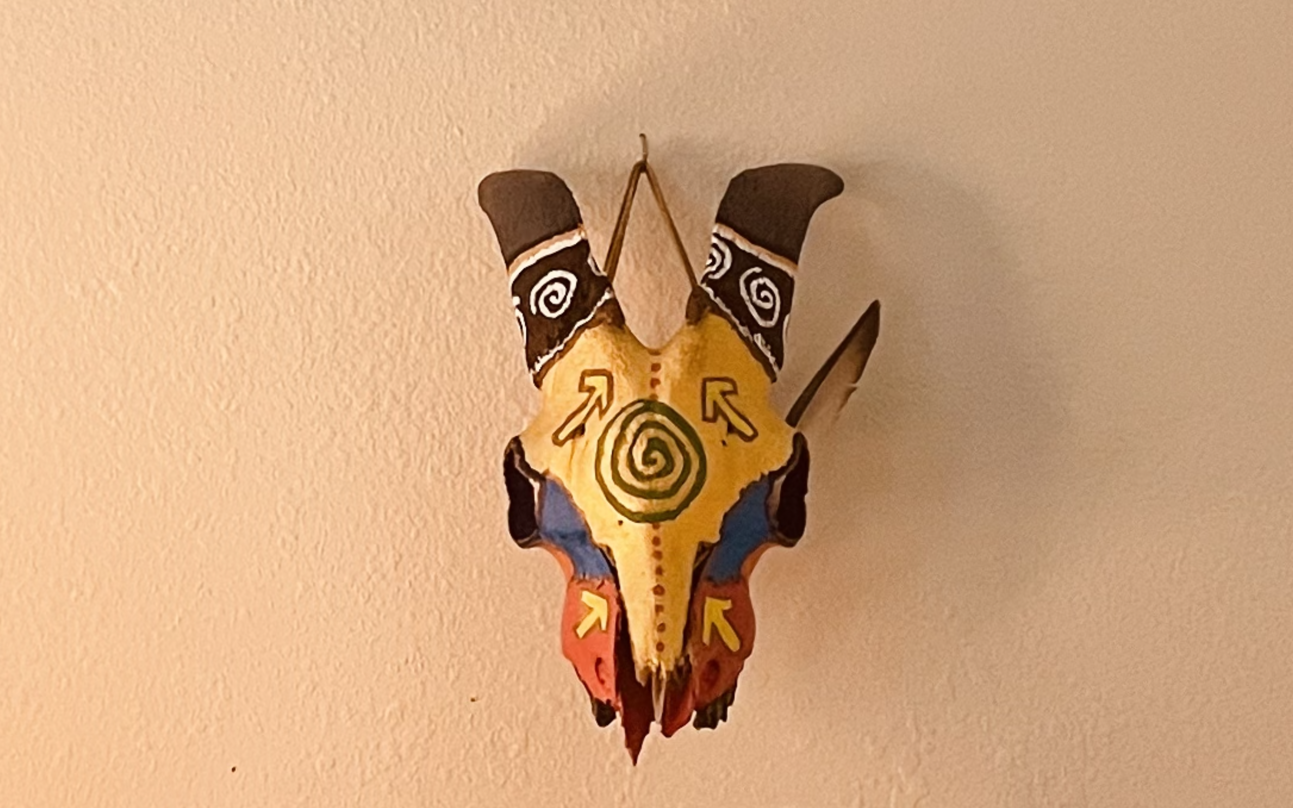 A painted goat skull with a feather stuck on the side; gold, pink, blue, brown and a light green pachamama symbol on the center of the forehead