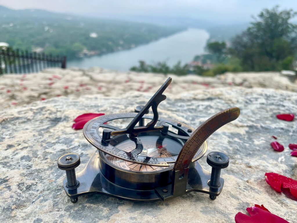 a sundial compass sitting on a rock cliff with rose petals around it; a river is in the background