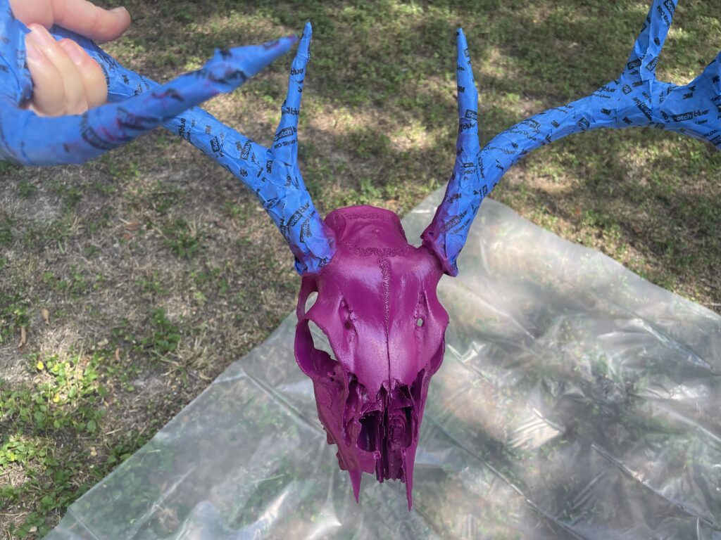 9-point deer skull with painters tape wrapped around the antlers. the skull has been spray painted a pinkish-purple color. a grassy yard and drop cloth are in the background
