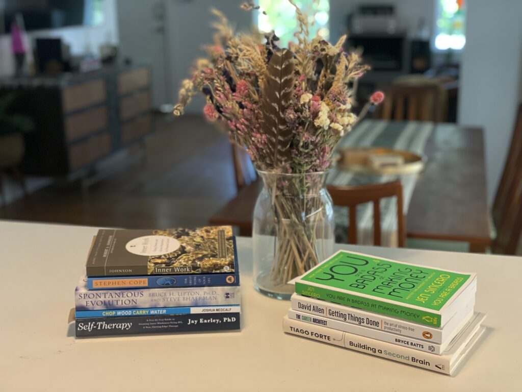 Two stacks of books sit next to a vase. Titles include: Inner Work, The Great Work of Your Life, Spontaneous Evolution, Chop Wood, Carry Water, Self-Therapy, You Are a Badass at Making Money, Getting Things Done, The Career Architect, and Building a Second Brain