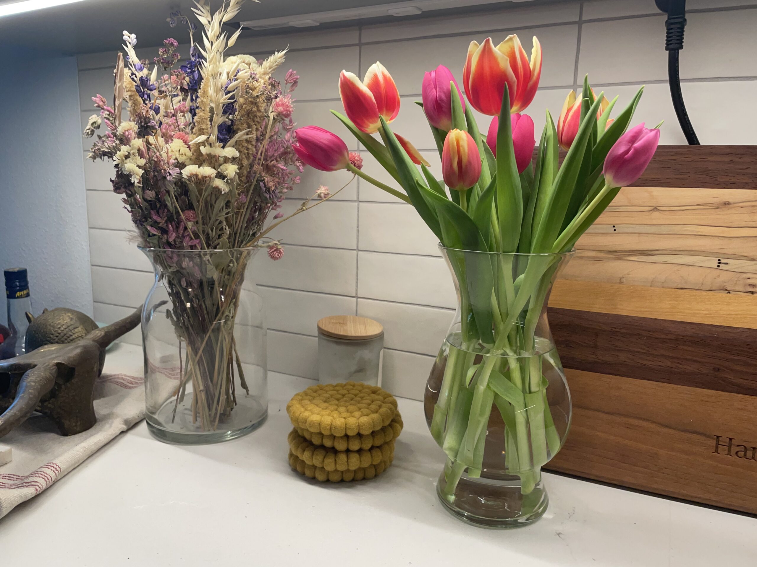 a vase of reddish orange and yellow tulips next to another vase of dried flowers and a stack of mustard yellow cloth coasters
