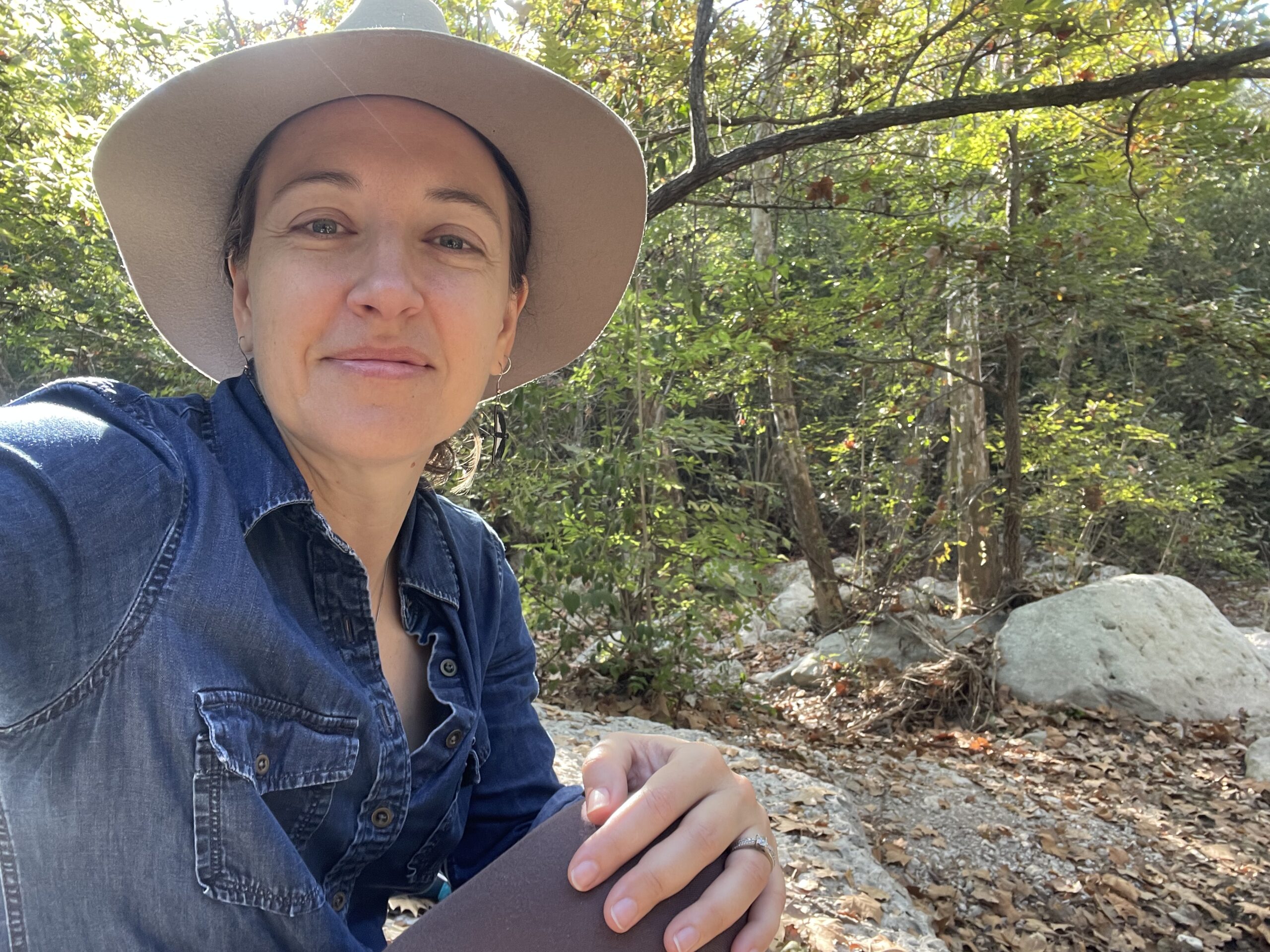 Amber wearing a brown felt hat and denim blue shirt, sitting outside with trees and rocks in the background