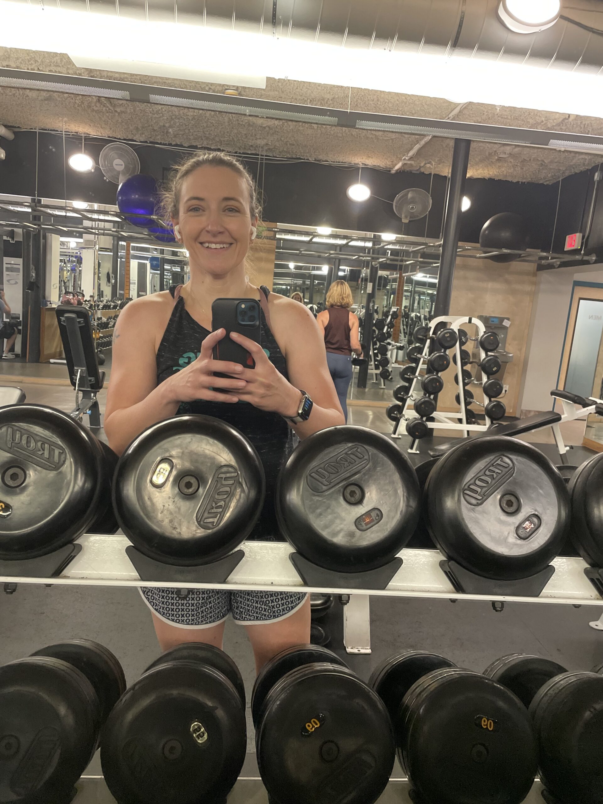 Amber at the gym taking a selfie in the mirror; dumbbells line the mirror in front of her