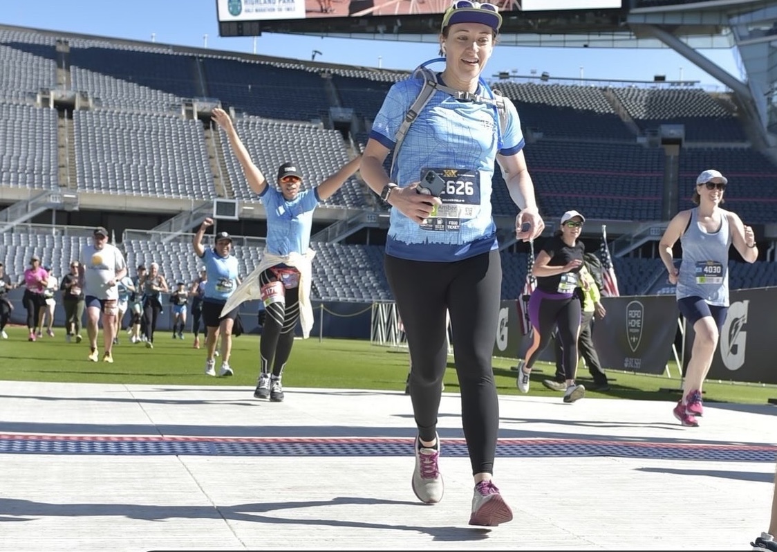 Amber running across the finish line; she is wearing black leggings and a blue t-shirt and camelbak hydration pack