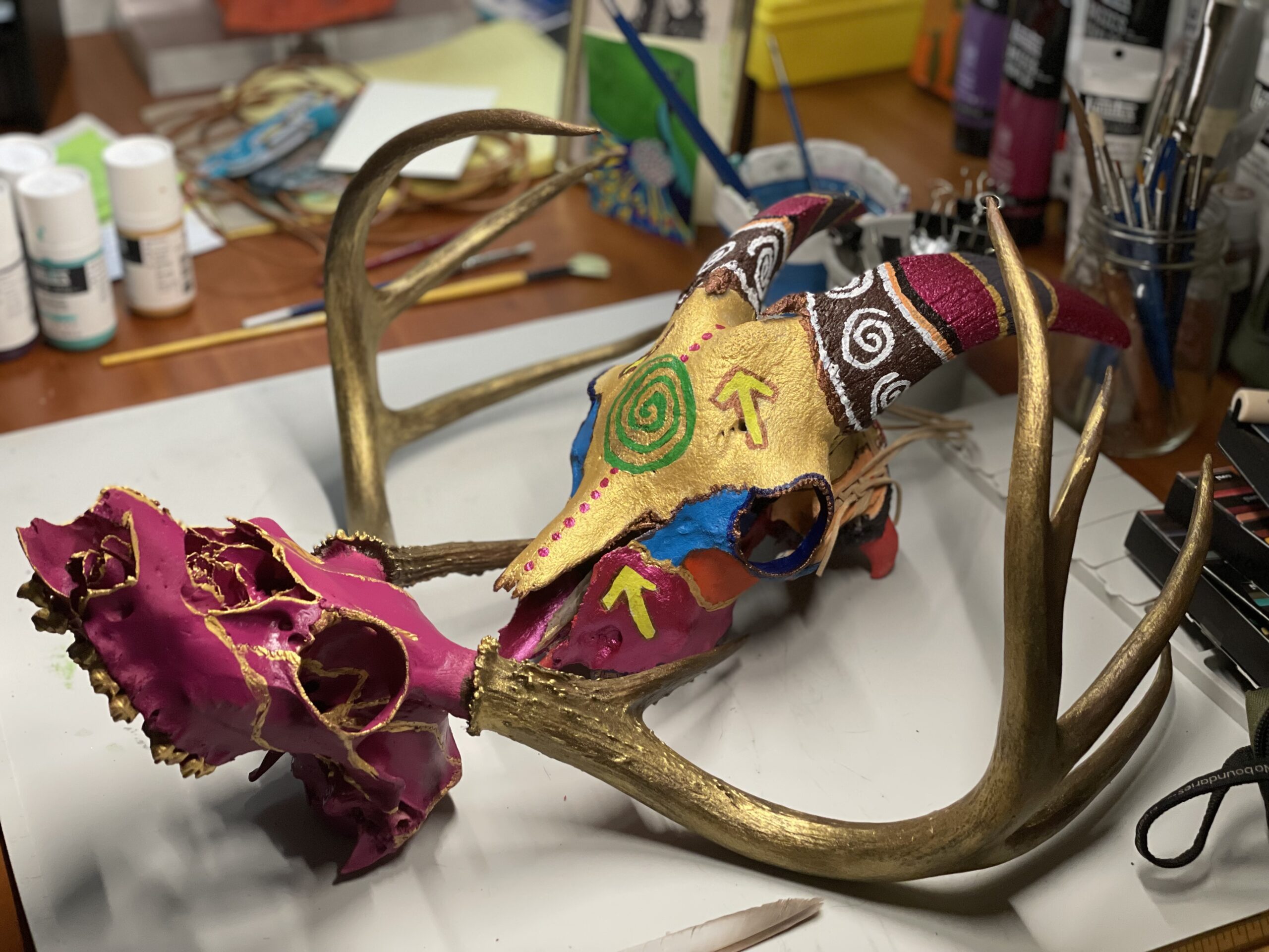 a painted deer skull next to a painted goat skull. The colors are bright pink and gold, with some green, blue, and yellow accent colors