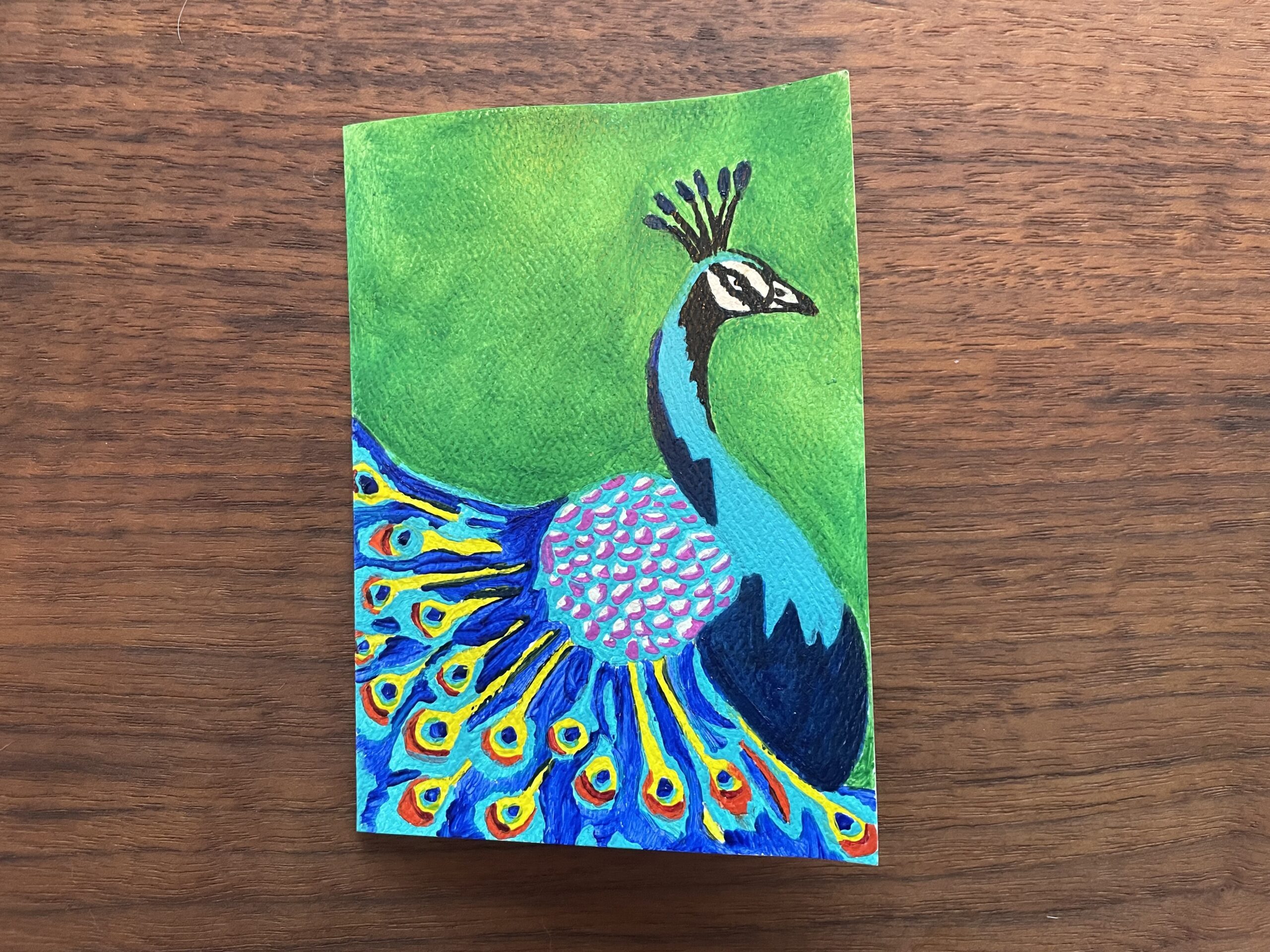 a small card with a painting of a colorful peacock on a green background