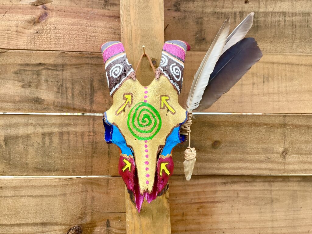 a colorfully painted goat skull with feathers and an arrowhead attached with leather strips