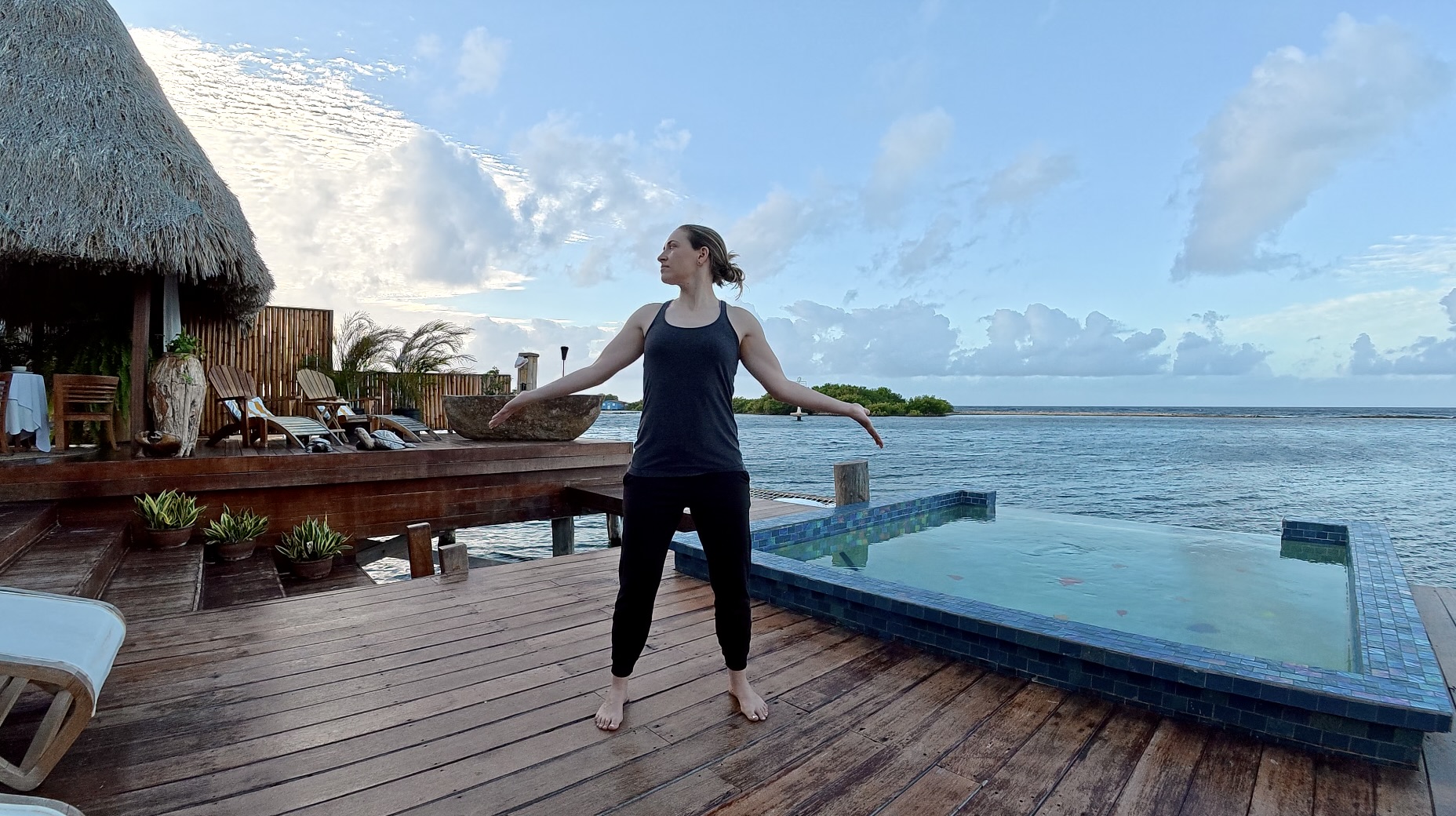a woman wearing a gray tanktop and black sweat pants is standing on a wooden deck turning her head to one side and opening her arms in the qigong pose called owl turns its head; the ocean and a small swimming pool are in the background