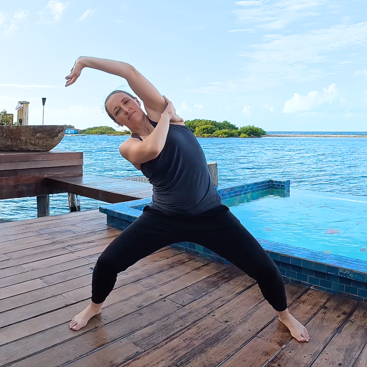 Amber is barefoot, wearing black pants and a gray tank top, standing in the Leopard qigong pose; a small pool and the ocean are in the background