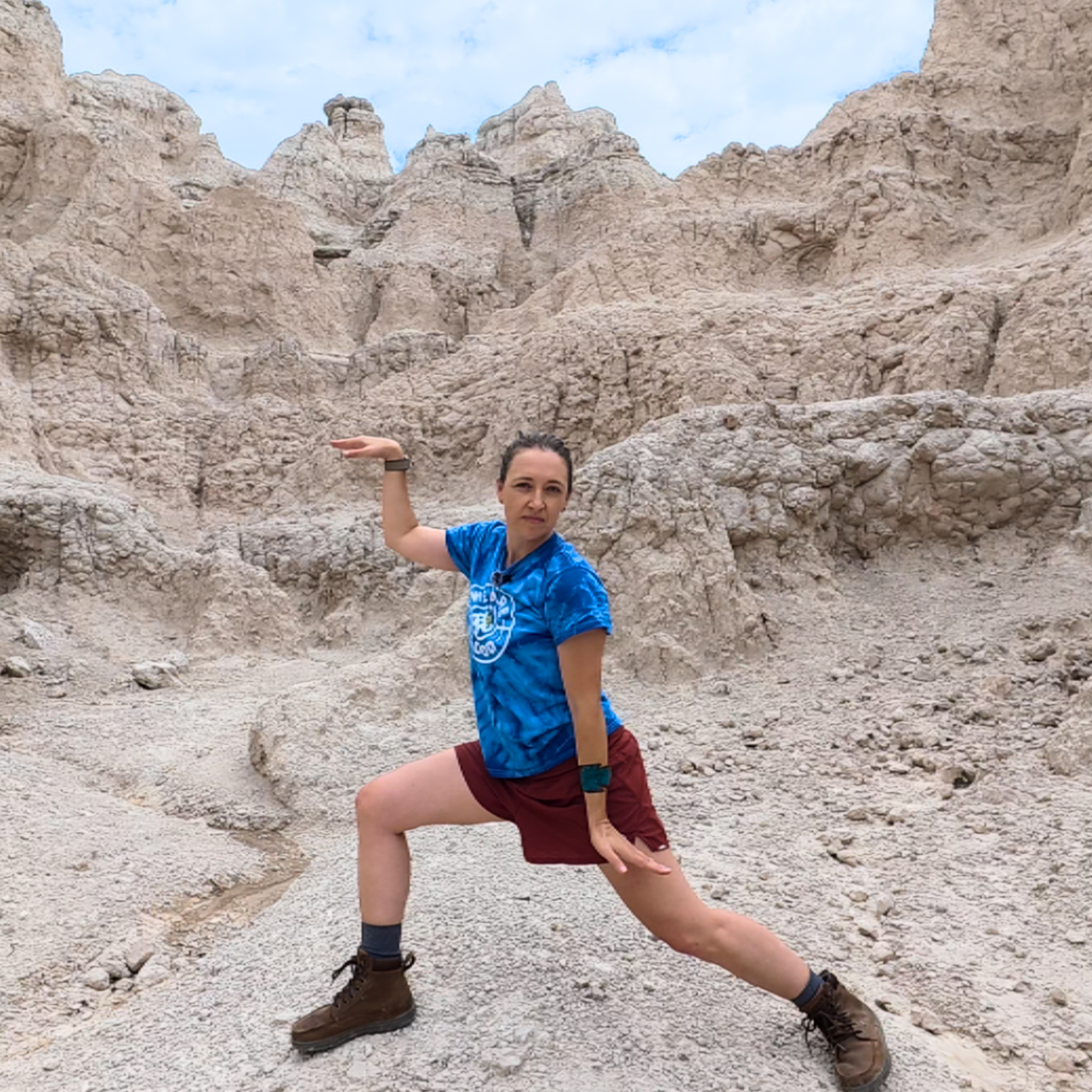 Amber wearing a blue tie dye T-shirt, maroon colored shorts, and hiking boots in the Snake qigong pose. Rocky, desert formations are in the background