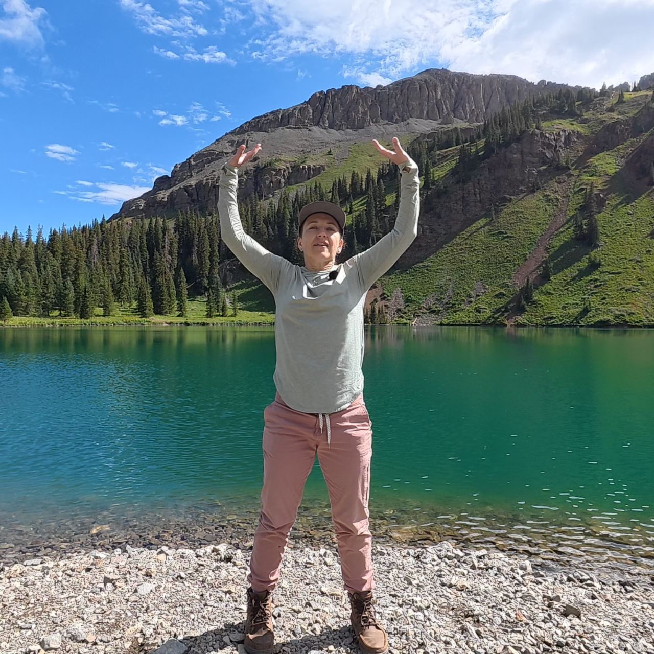 Amber smiling and standing with hands above her head; she is wearing a green shirt and pink pants; the background is a beautiful blue-green lake with a mountain behind it