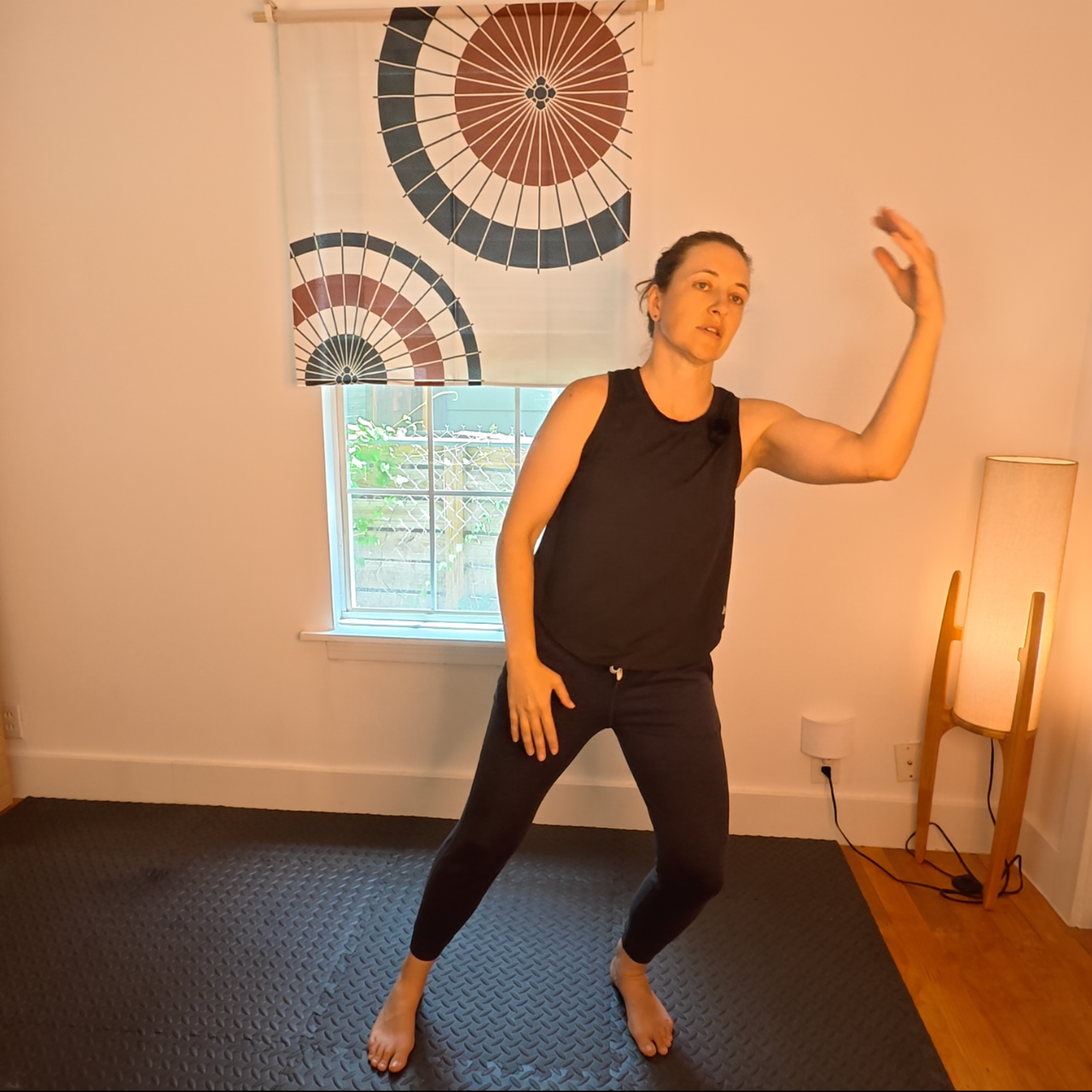 Amber wearing a black tank top and black sweatpants, moving in a qigong flow. The background has a window with a Japanese noren covering