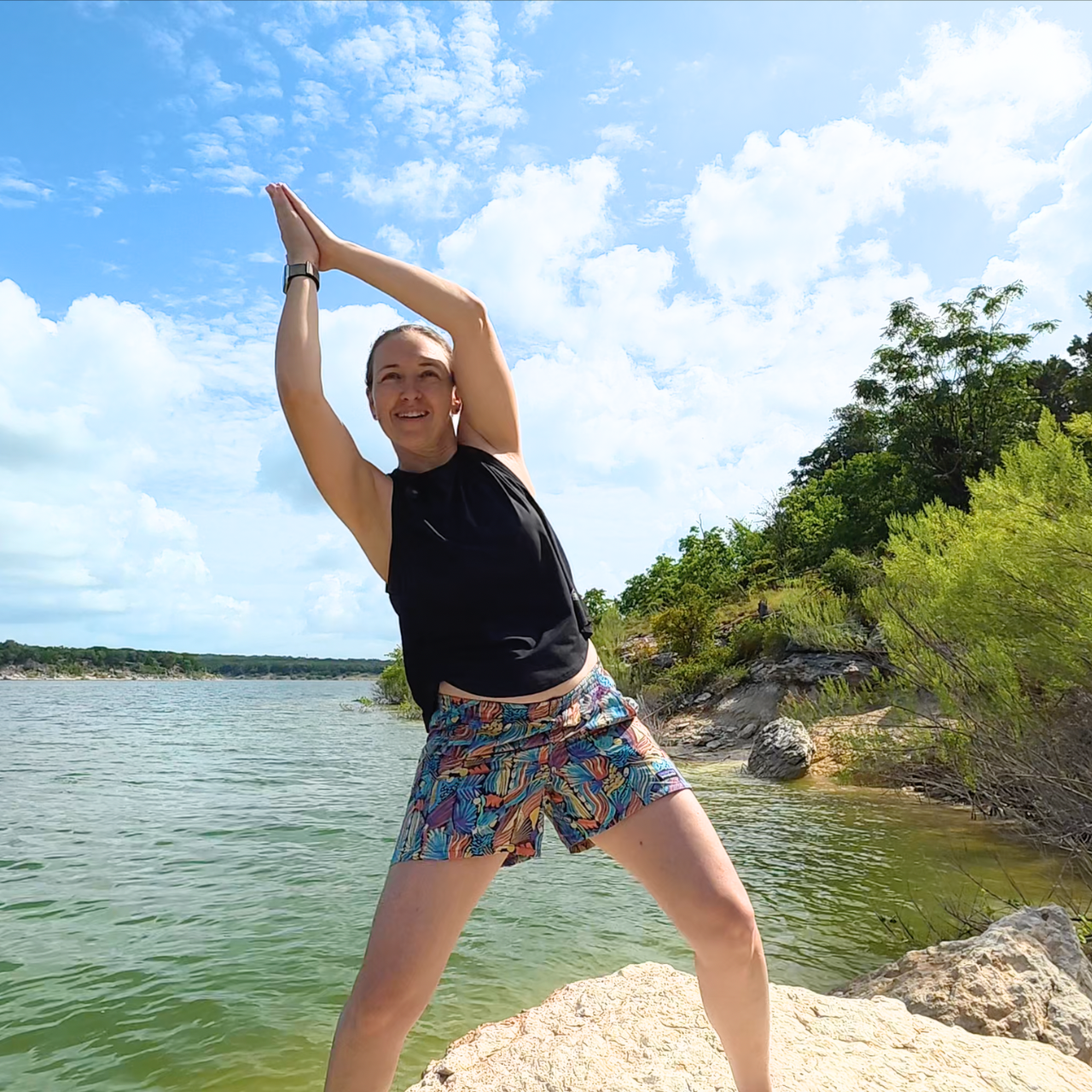 Amber is wearing a black tank top and patterned shorts, holding her hands above her head in a qigong pose while smiling