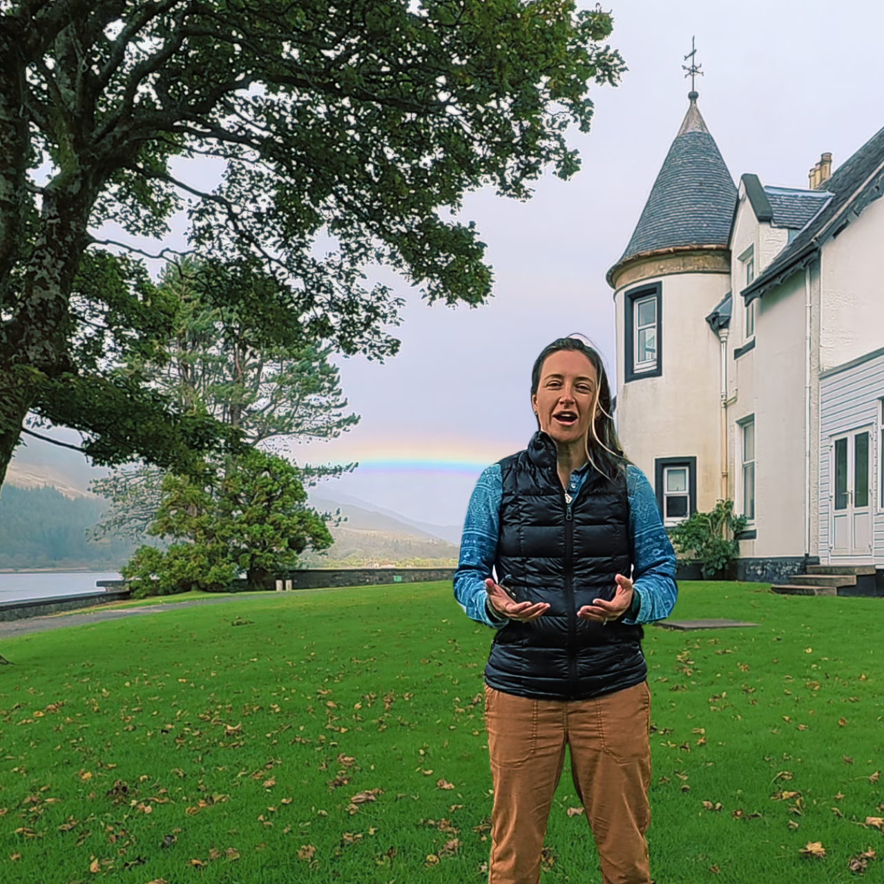 A woman in a black puffy vest, blue long-sleeve shirt, and brown pants stands on a bright green lawn. Background includes a white manor house, a rainbow, and a big tree by the lake